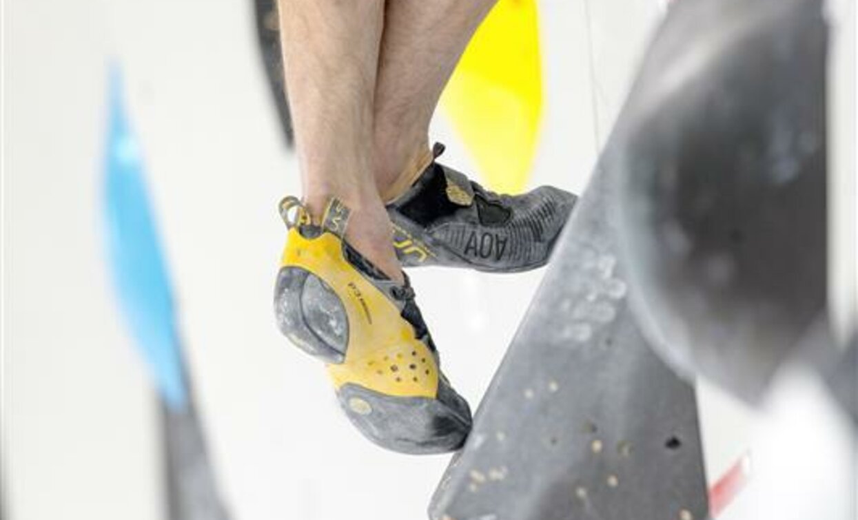 Un climber si appresta a scalare una parete artificiale. I suoi piedi indossano scarpette da arrampicata con suole colorate.