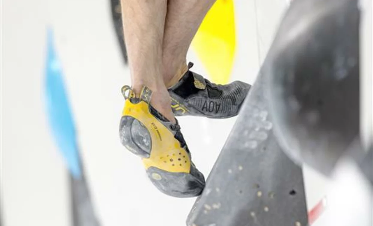 Un climber si appresta a scalare una parete artificiale. I suoi piedi indossano scarpette da arrampicata con suole colorate.