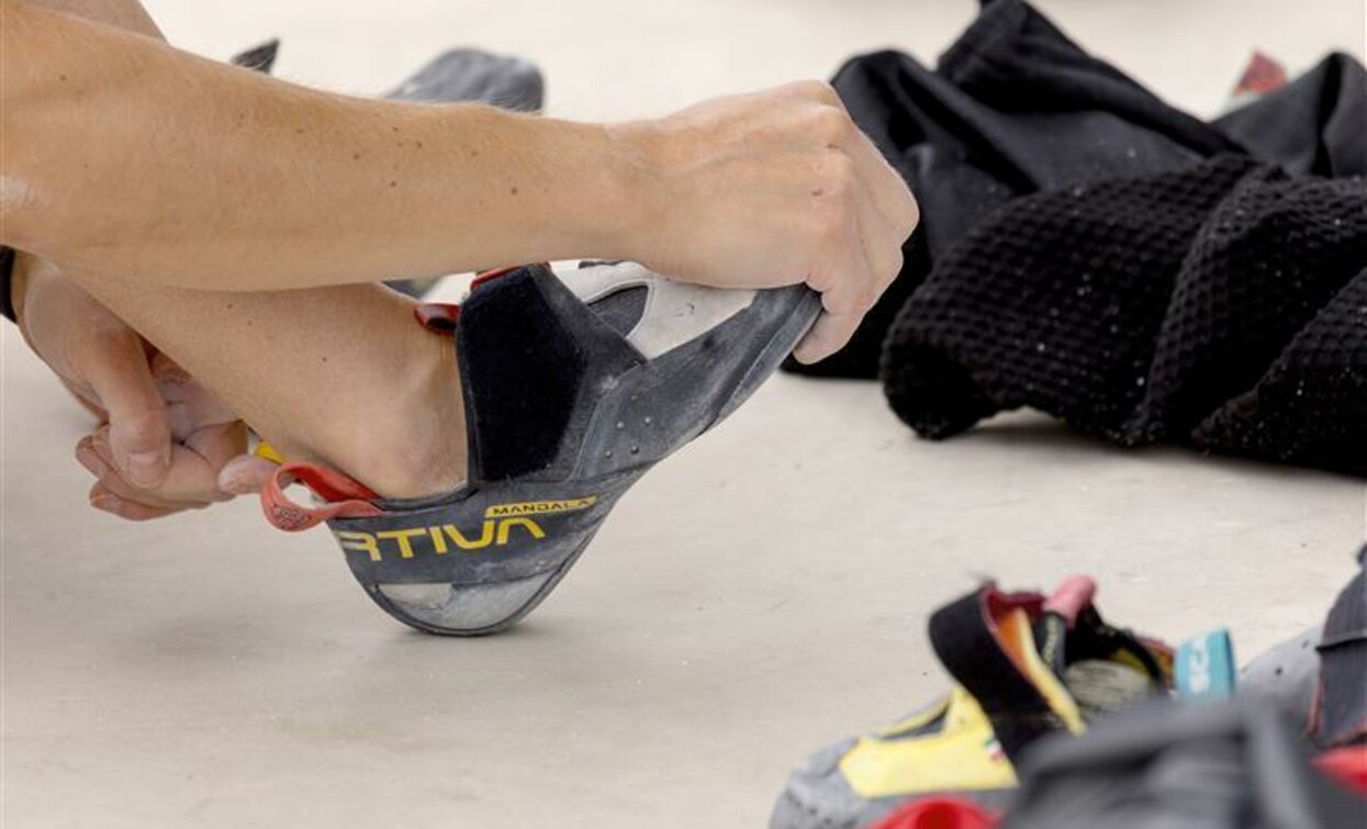 Una persona indossa una scarpetta da arrampicata. Sullo sfondo ci sono altre scarpe e attrezzature per l'arrampicata.
